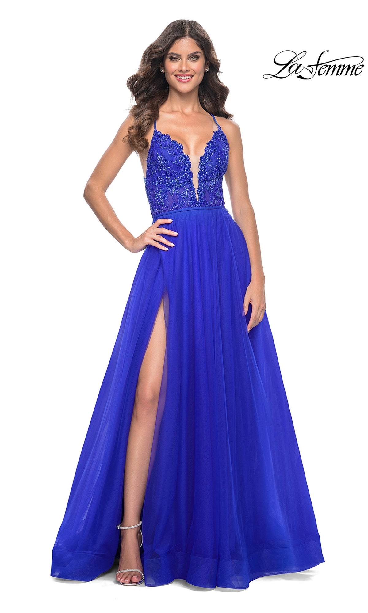 A woman in the La Femme Deep V-Neck Long Tulle Prom Gown 32059, featuring a sleeveless royal blue lace bodice, deep V-neck, high slit, and elegant a-line tulle skirt, smiles against a white background.